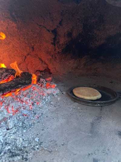 The image captures the interior of a rustic, wood-fired oven or hearth, viewed from an angled perspective looking downwards and into the chamber. The walls of the oven are rough and textured, appearing to be made of stone or clay, with areas of dark charring and soot towards the upper left, transitioning to lighter, ash-covered surfaces on the right.

The focal point of the image is a round, flatbread-like item, light brown or beige in color with a slightly textured surface, resting inside a dark, possibly cast-iron, pan or dish. This pan is set against or embedded into the right-hand wall of the oven, above the fire.

Below the pan and extending towards the bottom and left of the frame, a fire is actively burning. Several pieces of wood are visible, some still glowing with orange and red flames, while others have been reduced to bright red-hot embers. A significant amount of light grey ash covers the floor of the oven, particularly on the right side, with many glowing embers interspersed within it. The vibrant reds and oranges of the fire and embers illuminate the scene, casting a warm glow on the surrounding oven walls and highlighting the texture of the bread and the rough surfaces of the oven. The overall impression is one of traditional cooking in a hot, smoky environment. No text is visible in the image.