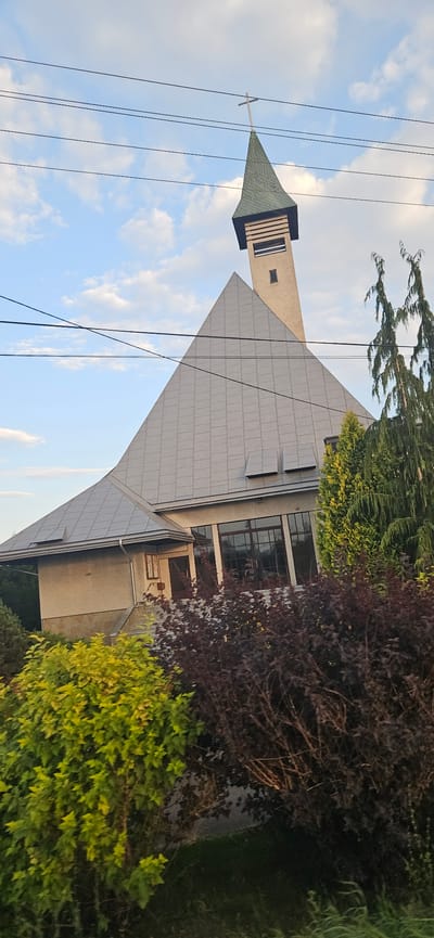 The image depicts a modern church with a prominent, tall, pointed steeple topped with a cross. The church building features a large, steeply pitched grey roof made of metal panels, creating a striking geometric design. The facade is light-colored, with a section of stone at the base. Large windows are visible, reflecting the sky and surrounding greenery. In front of the church, dense bushes with yellow-green and dark red foliage are growing, partially obscuring the lower part of the building. The scene is set against a bright blue sky with scattered white clouds. Several dark wires are stretched across the upper portion of the image, suggesting the presence of power lines. The overall impression is of a contemporary religious building situated in a somewhat natural or suburban environment. The image is captured from a slightly tilted perspective. Based on the provided context, this scene is located in the city of Iskrzynia, Poland.