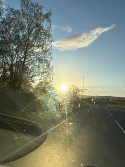 The image shows a view through a dirty car windshield towards a landscape with a blue sky and a wispy white cloud in the upper left. Below the cloud is a leafy tree branch. In the center, the sun shines brightly, obscured by foliage and the dirty windshield. On the right side of the image, a road with a traffic light is visible, though the details are blurred due to the windshield's condition. The setting appears to be an outdoor environment on a sunny day, possibly in Oswestry, United Kingdom, given the request. No people or specific actions are visible. The reflection of the car's interior is visible in the lower right corner.