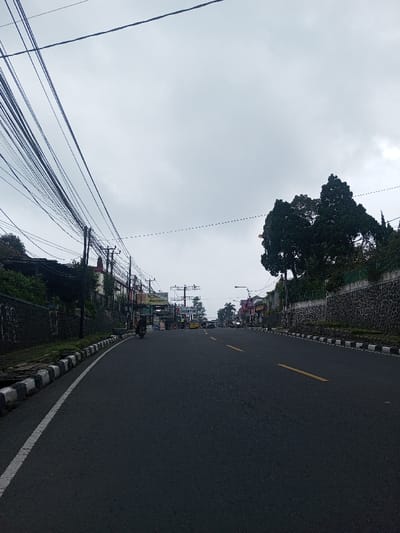 An overcast day casts a muted light on a straight, asphalt road marked with a single yellow line. Power lines tangle against the gray sky. A motorcycle travels in the distance. Buildings and stone walls line the sides of the road, flanked by greenery. White and black painted curbs border the road's edge. The perspective creates a converging effect, drawing the eye toward the horizon.