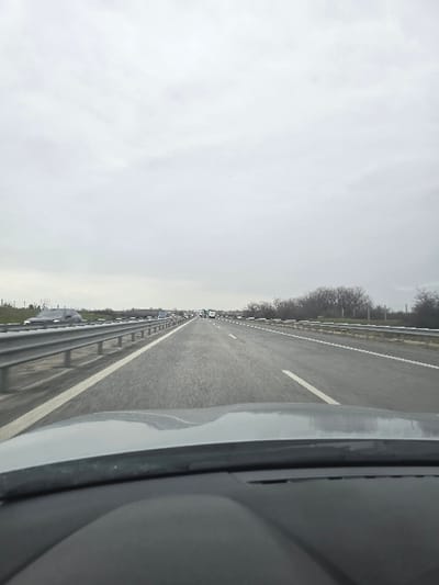 The image depicts a multi-lane highway scene in the city of Svoboda, Bulgaria, captured from inside a vehicle looking forward through the windshield. The vehicle's dashboard is visible across the bottom of the frame. The highway ahead features at least two lanes of travel in the same direction, delineated by white dashed and solid lines on a dark asphalt surface. Metal guardrails line both the left and right sides of the visible roadway. In the mid-to-far distance, traffic appears congested or slow-moving, with multiple vehicles including passenger cars and at least one white semi-truck visible in the rightmost lane. A silver-colored sedan is partially visible in motion in the left lane in the mid-ground. The sky is uniformly overcast and grey. The right side of the highway is bordered by a strip of bare deciduous trees and shrubs beyond the guardrail, while the left side shows open ground also separated by a guardrail.
