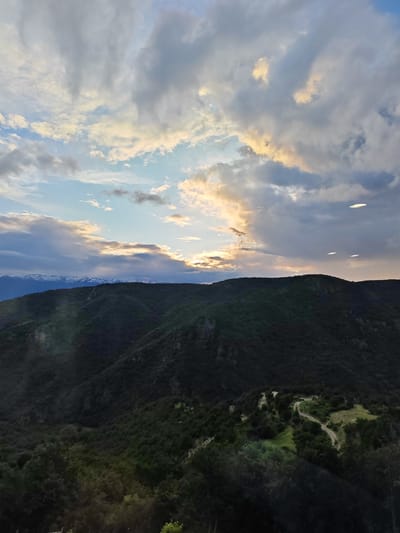 The image presents a panoramic mountainous landscape at either sunset or sunrise. The sky occupies the majority of the frame, featuring a dynamic interplay of clouds ranging from wispy to dense, in shades of blue, grey, and golden-orange. A bright, warm glow from the low sun pierces through the clouds along the horizon, creating a dramatic contrast.
In the midground, successive layers of densely forested mountains dominate, appearing dark green, almost silhouetted, against the brighter sky due to the low light. To the far left on the horizon, a line of distinct snow-capped peaks is visible, indicating a distant, higher mountain range. The lower right portion of the image reveals a clearer, lighter green area within the forest, where a winding dirt road or path can be seen traversing the terrain.
Four bright, elliptical light artifacts, likely lens flares or reflections, are present in the upper right quadrant of the sky. The scene evokes a sense of natural grandeur and tranquility. There are no people or discernible text visible in the image. The setting is a natural landscape in Leshten, Bulgaria.