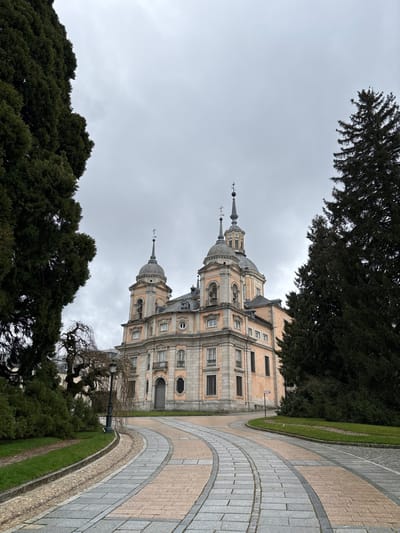 The image displays the Collegiate Church of the Holy Trinity (Colegiata de la Santísima Trinidad) at the Royal Palace of La Granja de San Ildefonso in Real Sitio de San Ildefonso, Spain, under a uniform overcast sky. The baroque-style building features a pale orange facade detailed with grey stone elements, including cornices, window surrounds, and pilasters. It is characterized by a prominent central dome surmounted by a lantern and a spire, flanked by two smaller domes also topped with spires, all culminating in crosses. Bells are visible within arched openings beneath the front domes. Numerous rectangular and arched windows are distributed across the structure, with balconies featuring iron railings. A primary entrance with large double doors is located centrally on the facade.
In the foreground, a curved paved pathway, constructed from alternating grey and light reddish-brown rectangular paving stones, leads toward the building. To the left of this path, a strip of green grass borders a cobblestone drainage channel. A tall, dense, dark green coniferous tree occupies the left edge of the frame, partially obscuring a leafless, gnarled tree and a black street lamp. On the right, a second tall, dark green coniferous tree partially obstructs the building. Green grass covers the grounds surrounding the path and trees. A small, reddish-brown directional signpost is partially visible on the grass to the right of the pathway. No individuals, animals, or vehicles are present within the scene.