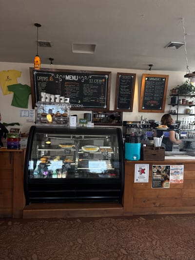The image captures the interior of a cafe named "Cafe Karma" in South Padre Island, United States of America. The perspective is from a patron's viewpoint, slightly angled. Several black chalkboards display the cafe's menu, with sections for "SANDWICHES," "CREPES," "DRINKS," and "ICE CREAM."

A glass-fronted display case contains various baked goods and desserts. A woman stands behind the counter, partially visible.

Posters and advertisements are affixed to the wooden wall behind the counter. These include "The Listening Sessions", "Solego Festival", and a "Best Coffeehouse" award.

T-shirts featuring the cafe's logo hang below the menu boards. A blue water dispenser sits on the counter. Various bottles and coffee-making equipment are placed on shelves above the counter. The floor is covered in a patterned brown rug.