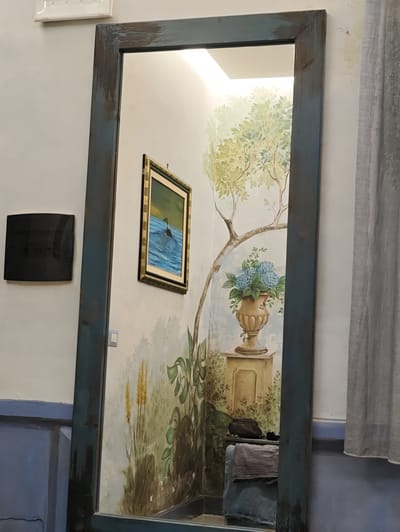 This is a full-length mirror with a distressed blue wooden frame, reflecting a room with painted murals. On the left wall of the reflection, a framed painting depicts a black bird or duck swimming in blue water under a yellow and green sky. To the right of this painting, a large black rectangular object is mounted on the wall. The background mural features a stylized tree with green foliage, a classical urn filled with blue hydrangeas, and a pedestal. Lower sections of the mural show green plants and yellow reeds. In the reflected foreground, there is a stack of striped fabric, a dark bag, and a blue upholstered stool. A grey curtain hangs on the right side of the frame. The scene suggests an indoor environment, possibly a home or a shop in Pozzuoli, Italy, given the context.