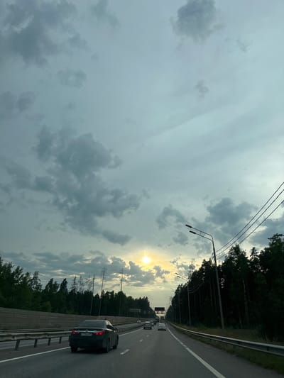 The image depicts a highway scene in Khlyupinskogo Lesn-Va, Russia, under a cloudy sky with the sun partially visible. A dark-colored sedan is traveling on the highway in the foreground, with its license plate, A775AB/50, clearly visible. Several other vehicles are visible further down the road. The highway is bordered by a sound barrier on the left and a dense forest on the right. Several streetlights line the right side of the road, and power lines run above the trees. The overall impression is of a scenic drive through a forested area on a day with variable weather.