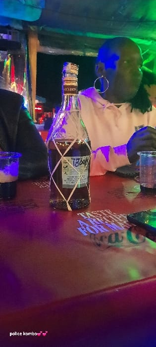 A bottle of Barcelo rum sits on a table. A person with round glasses and dark hair stands behind the bottle, wearing a light-colored top. The scene is illuminated by blue and green lights, creating a vibrant and colorful atmosphere. A plastic cup is visible in front of the bottle. The table appears to have writing on it, partly obscured by the bottle.