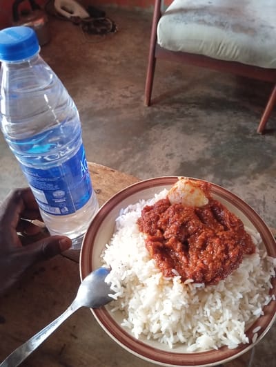 The scene depicts an indoor environment in Okere-Urhobo, Nigeria, presenting a meal and drink on a wooden surface. A round bowl with a white interior and brown rim contains a serving of white rice, topped with a substantial portion of reddish-brown stew or sauce. Embedded within the stew is a piece of a boiled egg, showing both white and orange yolk. A silver-colored spoon is positioned with its bowl resting in the rice on the left side of the dish. Adjacent to the bowl, a clear plastic bottle of Aqua-branded water with a blue cap is held by a dark-skinned left hand. The bottle is approximately three-quarters full. The background reveals a light grey concrete floor. In the upper left background, a metallic kettle and several dark, indistinct objects are visible on the floor. To the right, a portion of a wooden chair with a light-colored, stained fabric seat cushion is partially visible.