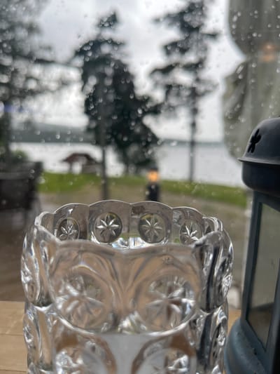 This is a close-up, slightly angled, eye-level shot of a clear glass votive holder in the foreground, with a blurred outdoor scene visible through a rain-streaked window. The votive holder is intricately patterned with starburst designs and has a scalloped rim. To the right of the votive holder, a dark metal lantern is partially visible.
The background, viewed through the window, is out of focus due to rain droplets on the glass and the shallow depth of field. It depicts a body of water, possibly a lake or river, with a small, dark-roofed gazebo or structure on the far bank. Trees with dark green foliage are also blurred in the background, suggesting an outdoor natural setting. The sky is overcast and grey.
The scene appears to be set on a wooden surface, likely a table or windowsill. There are no people or animals visible. The overall impression is one of a quiet, possibly melancholic, rainy day observed from indoors. The location is not specified in the image or context.