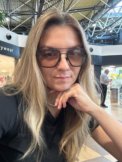 In Перепечино, Russia, a woman with blonde hair and glasses is taking a selfie. She is wearing a black shirt and a pearl necklace. Her right hand is resting on her chin, and she is wearing a ring on her right index finger. In the background, a man in a light blue shirt and black pants is standing and looking at a yellow object. The environment appears to be an indoor public space with a high ceiling and exposed metal beams.
