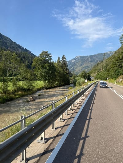 The image captures a scenic view of a mountain road in the vicinity of Predazzo, Italy. A paved road with white lane markings runs parallel to a guardrail on the right side of the frame. A dark gray car is driving on the road in the distance, followed by another white car. To the left of the road, a river flows with muddy water, bordered by lush green vegetation and trees. Beyond the river, a grassy field is visible, leading to a dense forest that covers the slopes of a mountain. In the background, majestic mountains with rocky peaks rise against a clear blue sky with scattered white clouds. The overall scene depicts a serene and natural landscape, typical of a mountain region.