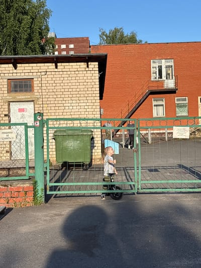 A young child, possibly a toddler, sits in a small, green ride-on toy with wheels, seemingly unattended, behind a green metal gate. The child is dressed in a light-colored dress with a pattern. The ride-on toy has a light blue seat. The gate is partially open, allowing a view of the child and the toy.
Behind the gate, to the left, is a building with light beige brickwork. There is a dark-colored vertical pipe running down the side of this building. A large, green trash bin with wheels is situated just inside the gate. Above the brick building, a red brick building with multiple windows and a fire escape is visible.
Further in the background, another red building can be seen, and trees with green foliage are present. The sky is a clear, bright blue, suggesting a sunny day. The ground surface appears to be asphalt. A large shadow is cast on the asphalt, likely from the gate or the adjacent buildings.
On the green gate, a sign is attached, and another sign is visible on a white metal panel below the gate. The overall scene appears to be an outdoor area next to residential or industrial buildings, possibly a courtyard or alleyway. The time of day seems to be midday due to the bright sunlight and distinct shadows. The emotion conveyed is one of stillness and a slightly eerie feeling due to the unattended child.
Visible text: A sign on the green gate, and a sign on a white metal panel near the bottom of the gate. The text on the white panel appears to be in Cyrillic script, with some characters visible like "МАШИНЫ" and "НЕ СТАВИТЬ !!!". Another sign above this reads "ТЕРРИТОРИЯ НАХОДИТСЯ ПОД ОХРАНОЙ ОБЪЕКТОВ И ПОД ВИДЕОНАБЛЮДЕНИЕМ ООО «Велес-Холдинг»".