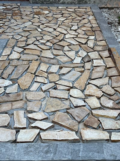 The image shows a close-up, overhead view of a stone pathway being constructed. The pathway is composed of irregular, flat pieces of stone in varying shades of tan, beige, and light brown, laid out in a seemingly random pattern with visible gray mortar filling the gaps between the stones. The stones have rough, natural edges. The pathway is bordered on the left and bottom by what appears to be a more uniform, gray stone slab or tile. The top and right edges of the pathway show glimpses of a darker, rougher surface, possibly soil or gravel. There are no people or animals visible in the image, and no identifiable landmarks or context suggesting a specific location within Menenkyovo, Bulgaria. The focus is entirely on the construction materials and pattern of the pathway.