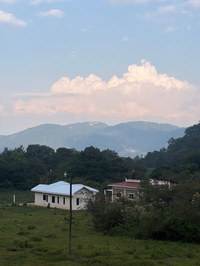 The scene depicts a rural landscape near La Esperanza, Honduras. In the foreground, there is a grassy field with a few houses. One house has a white metal roof and light-colored walls, while another is brick with a red roof. A utility pole with a streetlight is visible near the white house. Beyond the houses and field, there is a dense forest. In the background, layered mountain ranges stretch into the distance, partially obscured by a large cloud formation. The sky is pale blue with scattered clouds.