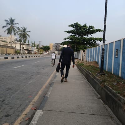 An outdoor, daytime scene captures an urban street in Crescent, Nigeria. The image, originally rotated, shows a paved road running from the foreground into the distance, bordered by a sidewalk on the right. A prominent black utility pole stands on the sidewalk towards the left.
The primary subject is a person, likely a man, dressed in a dark suit, walking on the sidewalk. Seen from behind, they carry a dark briefcase or bag in their left hand and wear brown shoes. Further down the road, another individual can be seen on what appears to be a bicycle or scooter.
To the right of the sidewalk, beyond a low curb, there is sparse ground vegetation leading to a blue-painted building wall with horizontal ridges. Several trees are visible, including a large leafy green tree behind the main subject and a couple of palm trees further along the street. The sky is bright and clear, indicating fair weather, likely late morning or afternoon. The overall atmosphere appears calm and routine, with people engaged in daily transit. No specific emotions are discernible, and no visible text is present.
