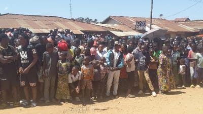 A large group of people, mostly dark-skinned, stand closely together outdoors on a dirt ground. A variety of clothing is visible. Buildings with corrugated metal roofs and a tall metal tower are in the background. The sky is blue, and the lighting suggests it is daytime.