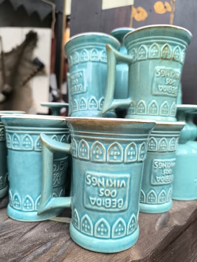 A close-up shot showcases a collection of turquoise mugs stacked on a wooden surface. The mugs are ornately decorated with embossed patterns, featuring a band of arched motifs near the rim and a rectangular panel with raised lettering. The text on the visible mugs reads "BEBIDA DOS VIKINGS" and "500" in reverse. One mug, positioned prominently in the foreground, is angled to reveal its handle and a partial inscription. The background is blurred, but hints of a shadowy figure and what appears to be a display rack suggest an outdoor market setting. The mugs are likely ceramic and exhibit a slightly aged appearance with subtle variations in color and texture. The scene is likely located in Óbidos, Portugal, a city known for its historic charm and artisanal crafts, and it is probable that these mugs are a souvenir item sold within the city.