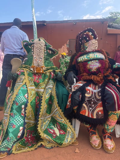 Two elaborately costumed individuals, likely participants in a festival or ritual, are seated outdoors in Porto-Novo, Benin. The individual on the left wears a green headdress adorned with shells and beads, a flowing green robe embellished with gold trim and sequins in patterns of animals and flowers, and holds a wooden object in their gloved hand. The individual on the right is similarly attired, with a headdress covered in shells and fabric, a mask-like face with colorful sequins and fringe, and a multi-patterned outfit featuring shells and sequins, including a beaded inscription that reads "JIKE MIJU" across the chest. They wear gloves and intricately decorated shoes. A man in a light blue shirt and jeans stands behind and to the left of the person on the left. In the background, a red-brown building with a corrugated metal roof is visible, along with a green pole with white lettering and other people in the distance, some wearing traditional clothing. The ground is a reddish-brown earth. The sky is clear and blue.