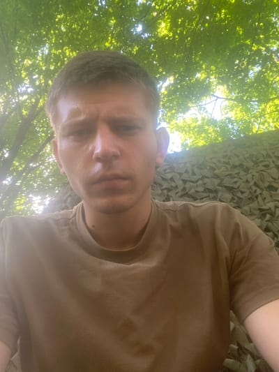 A young man with short brown hair and fair skin is pictured in a close-up, low-angle selfie taken outdoors. He is wearing a plain brown t-shirt. His expression is neutral, and his gaze is directed towards the camera.
The background features lush green tree leaves illuminated by sunlight from above. To the right of the man, partially visible, is a camouflage netting with a geometric pattern, suggesting an outdoor or possibly military setting. The environment appears natural, with the presence of trees and foliage. No specific landmarks or buildings are visible that would indicate a precise location within Shelkovo, Russia. The lighting suggests it is daytime, likely late morning or afternoon due to the dappled sunlight filtering through the leaves.