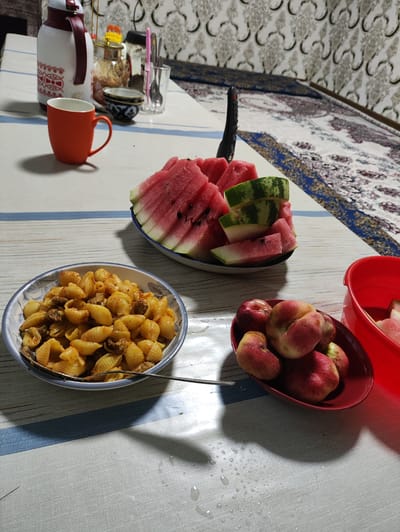 The image shows a table set for a meal in a room that appears to be in Navruz, Uzbekistan. On the table, there is a bowl of pasta with meat sauce, a platter of sliced watermelon, and a bowl of peaches. There is also a thermos, a mug, and some cutlery on the table. The table is covered with a striped tablecloth, and the background shows a wall with a patterned fabric hanging on it. The scene suggests a casual, home-style meal, possibly a picnic or a gathering. The presence of traditional Uzbek elements in the decor and food suggests a cultural context. The lighting suggests it is daytime, with some natural light filtering in.