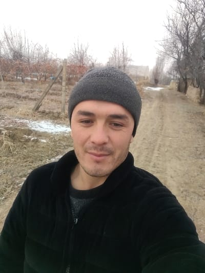 A man with a gray beanie and a black jacket stands outside. He is fair-skinned with dark hair and a slight smile. The background shows a dirt path leading into the distance, flanked by bare trees and low, straw-like vegetation. Patches of snow dot the landscape. The sky is overcast, creating a muted, gray light. A wooden fence stands to the left, adding depth to the rural scene.