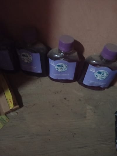 The image displays four bottles of "LaCoco Liqueur" standing upright on a gritty surface, possibly dirt or sand. The bottles have purple caps and labels featuring a circular logo with a depiction of what appears to be a wild animal, possibly a dog or wolf, and text. The background is a plain, light-colored wall. The lighting is dim, and the image quality is somewhat blurry, suggesting it was taken indoors with poor lighting conditions. There are no people visible in the image, and no discernible activities or interactions are taking place. The scene is static and appears to be a simple arrangement of the bottles. The location within Jalingo, Nigeria, cannot be determined from the visual information provided in the image.