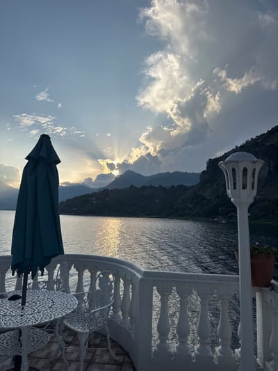 A serene landscape features a body of water reflecting sunlight, bordered by mountains and a partly cloudy sky. In the foreground, a closed blue umbrella stands beside ornate white metal patio furniture on a tiled terrace, enclosed by a white balustrade and decorative lamp. The setting conveys a tranquil outdoor ambiance, potentially overlooking a lake or sea from a high vantage point.
