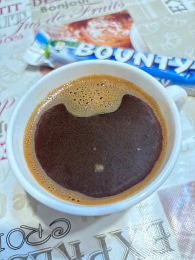 The image shows a close-up overhead view of a white mug filled with dark coffee, topped with a layer of foam. The coffee has a rich brown color and the foam is a lighter tan with small bubbles. A wrapper for a "Bounty" candy bar is visible in the background, partially obscuring the mug and resting on a patterned surface. The surface appears to be a tablecloth or placemat with various words and decorative elements, including some French text and ornate lettering. The lighting suggests it is indoors, possibly in a home or cafe setting. There are no people or animals in the image. The location in the city of Zubov, Russia is not determinable from the visual information provided in the image.