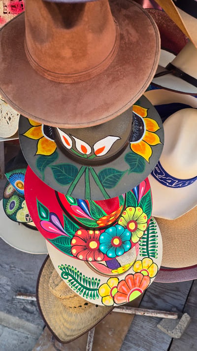 A stack of artisan hats, likely from a street vendor in Puerto Nuevo, Mexico, are displayed. The hats are of various styles and colors, with some featuring vibrant, hand-painted floral designs. The top hat is a plain brown, possibly made of felt, while the hats below are adorned with colorful depictions of sunflowers, other flowers, and leaves. The hats are arranged in a staggered fashion, creating a visually rich and textured composition. The background appears to be an outdoor setting, with hints of wooden surfaces and possibly other market stalls. The lighting suggests it is daytime, with natural light illuminating the hats.
