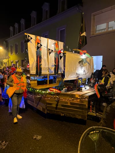 This image captures a nighttime scene on a wet asphalt street in the city of Wasserbillig, Luxembourg, depicting a pirate-themed parade float and associated participants.

The central subject is a multi-component float constructed to resemble a wooden boat or ship. The lower section consists of dark wooden planks, decorated with a string of warm-toned miniature lights. Above this, a platform supports several vertical white fabric banners. Each banner features a large, colorful parrot amidst tropical foliage. Between these, smaller black pennants with white skull and crossbones motifs are strung. On the right side of the float, a larger white banner displays the text "CAPTAIN SIDE PIRATES" with a stylized emblem. A black flag with a white Jolly Roger design (skull and crossbones) is prominently displayed from a mast-like structure at the rear right of the float. Illuminated, decorative parrot figures are visible on the top edges of the banners. On the float's deck, a blue glowing object is visible beneath the banners, and a digital display showing "18" is present next to a dark box, possibly a power unit or fog machine. A red basket is also visible on the deck.

To the immediate left of the float, a male adult walks on the wet street. He wears a yellow hard hat, an orange high-visibility vest over a grey puffer jacket and grey pants, and light yellow sneakers. A multi-colored feather boa (yellow, orange, red) is draped over his left shoulder, and he carries a yellow bag on his left side. Blue, translucent plastic material trails behind his legs, suggesting part of a costume.

On the right side of the float, several individuals are visible. One female adult wears a yellow high-visibility vest over a dark jacket. Another female adult wears a white puffer jacket, and a male adult in a dark jacket stands behind her. These individuals appear to be associated with the float or part of the procession.

The background features multi-story buildings lining the street. The building directly behind the float is light green with multiple windows and decorative dormers along the roofline. A partially visible sign reading "THEISEN" is present on the lower left of this building. To its right, a plain, lighter-colored building has a window with a closed beige roller shutter. The street is dark and wet, reflecting the artificial lights from the float, a streetlight visible on the far left, and interior building lights. Two flat, dark rectangular objects are visible on the wet asphalt in the immediate foreground.