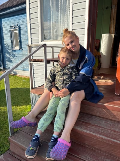 The image depicts a young woman with red hair pulled up in a bun, wearing a blue and navy jacket over a light-colored shirt, and a young boy in a camouflage jacket and light green pants with a pattern of cartoon vehicles. They are sitting on the wooden steps of a porch. The woman has her arm around the boy, and they are both looking towards the camera. The background shows a blue wooden house with white trim and windows, and a green door. A grassy area is visible to the left. The overall scene suggests a casual, outdoor setting, likely in a rural or suburban area, possibly a dacha or country house, given the architectural style of the building and the presence of a lawn. The location is specified as Dunilovo, Russia.