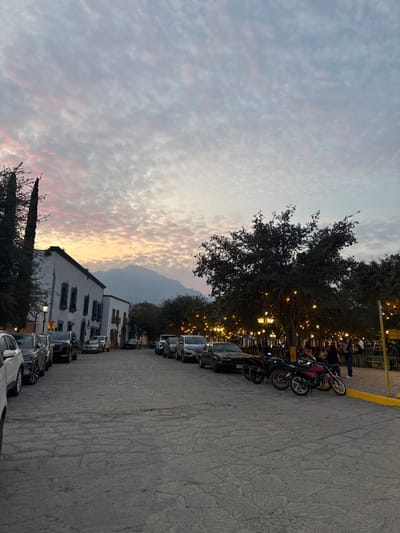 A streetscape with parked cars and motorcycles lines the right side of the image. The left reveals a cloudy sky with hints of orange, likely from a sunset. A building with white walls and multiple windows stands along the street. In the background, a mountain range is visible. Trees with string lights adorn the area, adding a warm glow. People can be seen walking near the lights, suggesting a public gathering space. The ground is paved with stone.