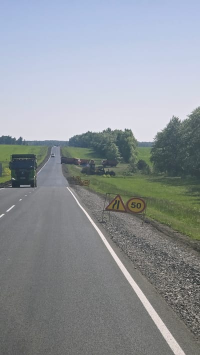 The image depicts a straight, paved road stretching into the distance under a clear, pale blue sky. The road has a white line marking the right edge. To the right of the road, there is a gravel shoulder with two traffic signs. The closest sign is a yellow triangle with a black border, pointing to the left, indicating a lane narrowing. Next to it is a circular sign with a red border and a white background, displaying the number "50" in black, signifying a speed limit of 50. Further down the road, on the grassy right side, are several construction vehicles including trucks and what appears to be a yellow bulldozer. Beyond the construction vehicles, a line of green trees forms a backdrop. On the left side of the road, a green truck is visible. The overall environment suggests a rural or semi-rural setting.
