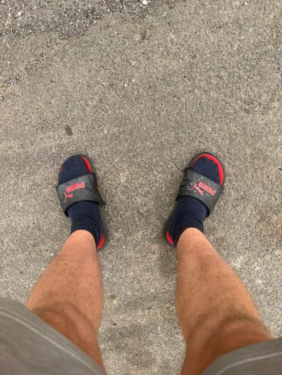 A first-person perspective shot shows a person's legs from the knees down, standing on a textured concrete surface with scattered gravel. The person is wearing black Puma slide sandals over dark navy socks. The sandals have a red trim and the Puma logo in red. The legs are hairy and tanned. The lower part of the frame is partially obscured by grey fabric, possibly shorts. The scene is an outdoor urban environment.