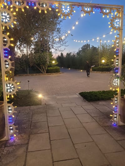 An evening outdoor scene captures a decorative archway adorned with numerous small lights in white, yellow, and blue, framing a paved pathway and surrounding garden. The archway's metallic structure features intricate, radial designs resembling ship wheels, each holding several lights. String lights stretch across the fading blue twilight sky, connecting to the archway and creating an overhead canopy of illumination.

In the middle distance, a person dressed in dark clothing and a cap walks away from the camera along the rectangular stone-tiled path. Lush green trees and bushes line both sides of the pathway, with additional warm light sources visible among the foliage and in the background. A stone wall runs along the right side of the paved area. The soft, artificial lighting creates a warm and inviting atmosphere as daylight transitions into dusk. The scene depicts a festive or pleasantly illuminated setting, likely a garden or courtyard, situated in Sogliano Cavour, Italy. No text is visible in the image.