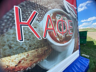 The image shows a close-up of a sign with text that appears to be in Cyrillic, with the letters "К" and "АФЕ" prominently displayed. Behind the text is a picture of a bowl of food, likely soup, and slices of bread. The sign is mounted on a blue corrugated metal structure and is outdoors, with green grass and a blue sky with clouds visible in the background. To the right of the sign, a green trash receptacle is visible. The overall scene suggests an exterior view of a cafe or food establishment. No people are visible in the image. The location of Yuldybayevo, Russia, is not explicitly indicated in the visual information provided.
