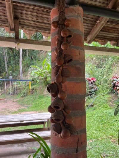 The image depicts a static, eye-level view of an outdoor covered area in Girón, Colombia, adjacent to a natural, vegetated landscape. A vertical, reddish-brown brick column with gray mortar lines occupies the central-right foreground, supporting an overhead wooden roof structure. A dark, fibrous rope or vine is wrapped vertically around this column, from which approximately two dozen dried natural objects are suspended. These objects include light brown, smooth, tear-drop or bulbous-shaped gourds and darker, textured, bell-shaped or truncated cone-shaped seed pods with hollow interiors.
In the lower-left midground, the top of a wooden picnic table with attached benches is partially visible. Beyond the table, a dirt or gravel surface transitions into a green grassy area. A blue and yellow metal swing set with two swings is positioned in the midground on this grassy expanse. The background on both sides of the column consists of dense, lush green vegetation, including numerous tall trees and various smaller plants, indicating a heavily vegetated environment. On the right side, a sloping green lawn displays clumps of plants with reddish foliage. No individuals, specific actions, or interactions are present in the scene.