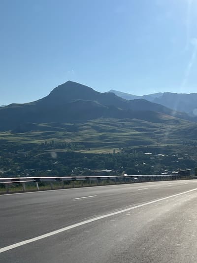 A wide, asphalt road with a white lane marker curves to the right in the foreground. A metal guardrail with black and white horizontal stripes lines the left side of the road. Beyond the road, a village is nestled in a valley. Houses with red and gray roofs are scattered amongst green trees and fields. Rolling hills covered in green fields and trees rise behind the village. A large, dark, mountainous landscape dominates the background, with a prominent peak in the center. The sky is a clear, bright blue with a slight haze in the upper right corner, suggesting a sunny day. The scene appears to be a rural landscape in or near Zangakatun, Armenia, given the mountainous terrain and village architecture.