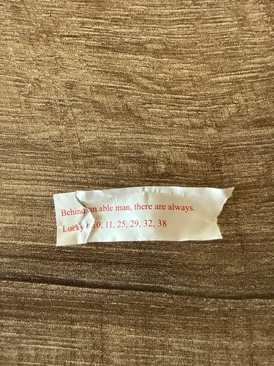 A close-up shot shows a partially torn fortune cookie slip resting on a textured, golden-brown surface. The text on the slip, printed vertically in red, reads: "Behind an able man, there are always." Below this, it states "Lucky # 10, 11, 25, 29, 32, 38." The slip is white with minor tears along the edges. No people or specific environmental elements indicating Darien, USA, are visible.