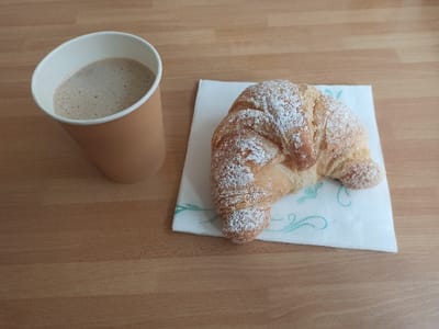 This scene, located in San Marzano sul Sarno, Italy, depicts a light-colored wooden surface, likely a tabletop. On the left, a plain, light brown paper cup contains a light brown liquid topped with a thin layer of beige foam. To the right, a crescent-shaped, golden-brown croissant, generously dusted with white powdered sugar, rests upon a white square paper napkin. The napkin features a light teal decorative swirl pattern in its lower portion. No individuals are present, and no actions or interactions are depicted.