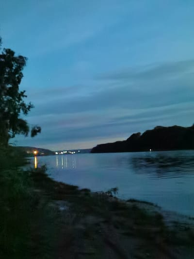 The image captures a twilight scene on the Kama River in Chaikovsky, Russia. The sky is a deep blue, with scattered clouds and a faint reflection of a celestial body visible in the upper portion of the frame. On the left bank of the river, a dark silhouette of trees and vegetation is present, with a bright yellow light source reflecting on the water. Across the river, the opposite bank features a dark, silhouetted hill or landmass. In the distance, a line of lights, likely from a settlement, is visible along the far shore, with their reflections shimmering on the water's surface. The water itself appears calm, with subtle ripples disturbing the reflections. The overall atmosphere is serene and tranquil, characteristic of dusk by a large body of water.