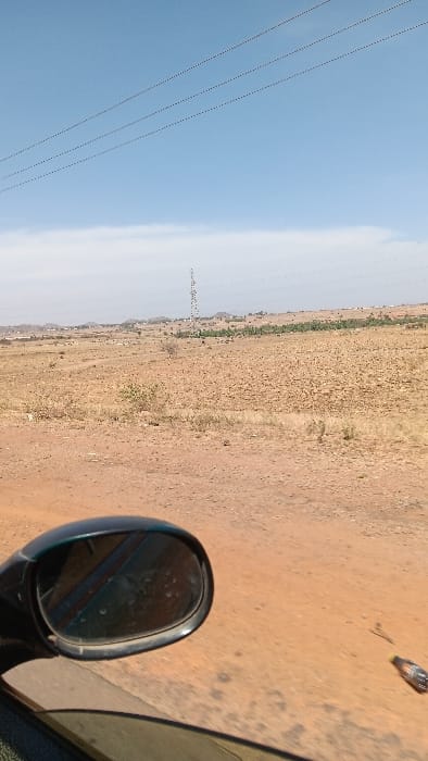 The image depicts a dry, open landscape in Barakin Tenti, Nigeria, viewed from the passenger side of a vehicle. The foreground shows a dusty, dark-colored automotive side mirror on the left and a portion of the vehicle's door and window frame below it. The immediate ground outside the vehicle is a reddish-brown dirt road, on which a small, clear plastic bottle with a yellow cap lies horizontally to the right of the mirror. The midground features expansive, arid terrain covered in yellowish-brown dirt and sparse, dry scrub vegetation. A tall, lattice-structured telecommunications or power transmission tower stands centrally in the mid-distance. Further back, a line of green foliage, possibly trees or crops, is visible, suggesting a more fertile area or distant settlement. The background consists of hazy, undulating hills or mountains stretching across the horizon. The sky above is a clear, bright blue, with three prominent, parallel overhead lines, likely power or communication cables, strung across the upper portion, populated by numerous small birds perched along them. No people or active interactions are visible.