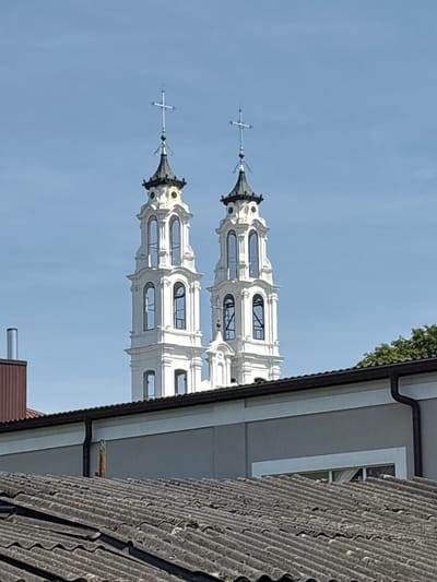 The image shows two white church towers with dark spires and blue crosses against a clear blue sky. The towers are ornate, with multiple levels and arched windows. In the foreground, a grey, tiled roof obscures the lower part of the church. To the left of the towers, a portion of a brown building with a metal chimney is visible. To the right, the tops of green trees and another building with a dark gutter are present. The architecture of the towers suggests they are part of a historic church, likely St. Michael's Archangel Church in Ashmyany, Belarus, given its characteristic twin towers. The scene appears to be a sunny day, with clear visibility.
