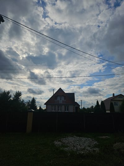 The image depicts a suburban residential scene in Gornye Moriny, Russia, under a cloudy sky. In the foreground, a dark, dense fence runs horizontally across the frame, partially obscuring the view of the houses behind it. To the left of the fence, a brick pillar stands out. The ground in front of the fence is covered in grass with some patches of scattered stones.

Behind the fence, two houses are visible. The house on the left is a two-story structure with a red roof, brick facade, and white window frames. It features a balcony on the upper level with a white railing. The house on the right has a brown roof and a lighter colored facade with visible window frames. Both houses appear to be residential properties with chimneys.

A line of trees, mostly dark green evergreens, is situated behind the fence and in front of the houses, suggesting a semi-rural or suburban setting. Overhead, multiple power lines crisscross the sky, adding a man-made element to the natural landscape. The sky itself is filled with dramatic, heavy clouds, with patches of blue sky peeking through, indicating potentially changing weather conditions. No people or animals are visible in the scene.