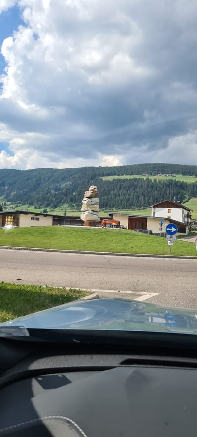 The image shows a view from a car in Toblach - Dobbiaco, Italy. In the foreground, the car's dashboard and windshield are visible. The middle ground features a grassy roundabout with a large, colorful sculpture of stacked rocks. Behind the roundabout, there are buildings, an orange excavator, and a road sign indicating a right turn. The background consists of a lush green, tree-covered mountain under a cloudy sky. The scene appears to be an outdoor public space.