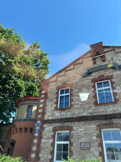 This media file, taken in Tartu, Estonia, captures an exterior view of a historic building on a bright, sunny day. The building's facade is a mix of rough-hewn light-colored stone and red brick, with distinct architectural details like stepped brickwork and decorative brick accents around the windows. Several windows with white frames are visible, reflecting the blue sky.
A prominent sign on the building displays the word "Aleksan" in dark lettering on a black banner, partially obscured by a yellow glow, suggesting it might be a historical marker or an old business name. Below this, a rectangular plaque is affixed to the stone wall. It contains text in Estonian, reading: "SELLE HOONE ASUS / AASTATEL 1880–1897 / TARTU / GAASIVABRIK". This indicates the building was once a gasworks.
Further down the wall, two blue street signs are visible: one reads "Aleksandri" and the other displays the number "42". A cylindrical section of the building, clad in light-colored wood and trimmed with red, is also visible, adding to the unique architectural character. Lush green trees with full foliage are positioned in the foreground and around the building, indicating it's a warm season, likely summer, given the vibrant greenery and clear blue sky. The overall impression is one of historical significance within a pleasant, natural environment. There are no people or ongoing activities depicted, focusing solely on the static architecture and its surroundings.