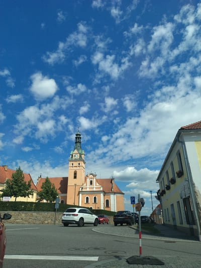 The image shows a street scene in Lhenice, Czechia, featuring a church with a clock tower as the dominant architectural element. The church is light orange with a green-roofed tower topped with a cross. To the right of the church is a yellow building with red-tiled roofs and flower boxes. In the foreground, a white SUV is driving on a paved road. Several other cars are parked or driving in the background. The sky is a bright blue with scattered white clouds. A stone wall with a hedge runs along the side of the road in front of the church. There are also various street signs visible, including a parking sign. The overall scene depicts a sunny day in a European town.