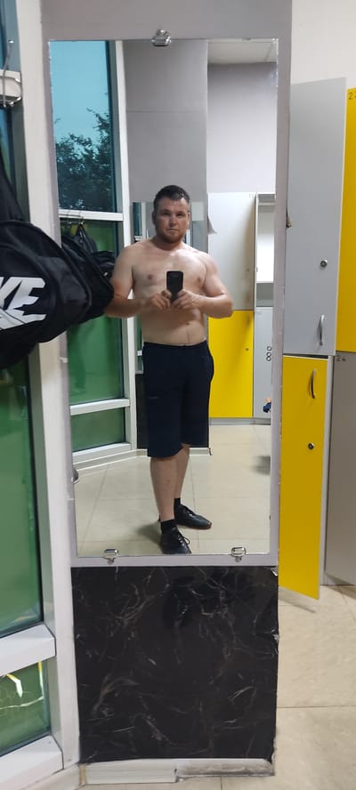 This media file shows a man taking a mirror selfie in what appears to be a locker room or gym. The man is shirtless, wearing dark athletic shorts and black athletic shoes. He is holding a smartphone in his right hand to take the picture. His reflection is visible in a large mirror that takes up a significant portion of the frame.

Behind the man's reflection, the locker room features several lockers. Some are grey and have silver handles and locks. Others are bright yellow and have black handles. The floor is a light-colored tile.

To the left of the mirror, there is a window with a greenish tint, through which trees and a bright blue sky are visible, suggesting it is daytime and the weather is clear.

To the right of the mirror is a section of black material with a marble-like pattern.

The man's expression is neutral as he looks at his phone. The overall impression is one of a casual moment captured in a fitness-related environment.

There is no visible text in the media file.