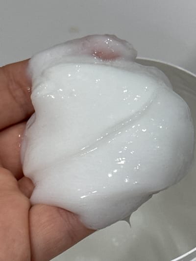 A close-up shot shows a person's hand holding a dollop of white, foamy product, possibly a cosmetic or cleaning substance. The product has a somewhat translucent appearance with small, reflective bubbles scattered throughout. The hand, visible from the fingers to the palm, appears to be a fair skin tone. The product is molded into soft, undulating shapes as it rests on the skin. The background is a plain, light gray surface, suggesting a clean, indoor environment. There is no visible text in the image. The lighting is soft and diffused, highlighting the texture and shine of the product without harsh shadows. The focus is entirely on the hand and the product, creating an intimate and detailed view of the material's consistency.