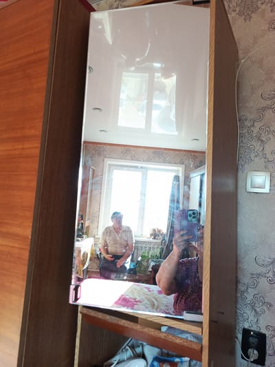 A medium shot captures a reflection in a tall, vertical mirror. The mirror is framed by a wooden cabinet with a wood-grain finish. In the reflection, a man with glasses and a light-colored collared shirt is seated in front of a window. To the right of the man, a woman is visible, holding a purple phone in her right hand, taking the photograph. She is wearing a purple and pink patterned top. The room appears to be a domestic setting, possibly a bedroom or living room, with patterned wallpaper on the right wall and a radiator visible behind the man. The floor in the reflection is partially covered by a rug. The scene is captured in Gusinoozyorsk, Russia.